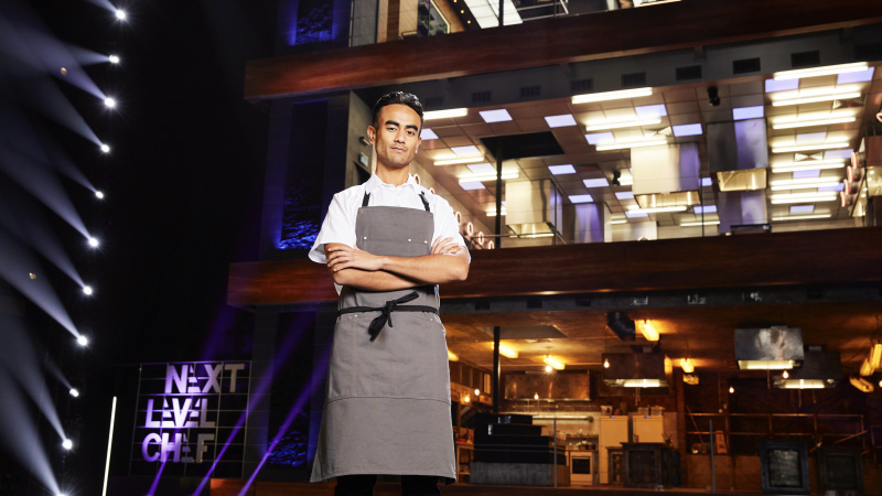   Ronan ผู้เข้าแข่งขันสวมเสื้อสีขาวและผ้ากันเปื้อนสีเทาอยู่หน้าห้องครัวใน Next level Chef