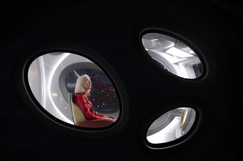Solos: Peg, interpretada por Helen Mirren, está sentada dentro de uma espaçonave vestindo um macacão vermelho. A câmera está do lado de fora, olhando através de três janelas em forma de ovo