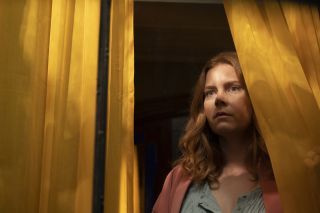 The Woman In The Window: Η Anna Fox (Amy Adams) στέκεται σε ένα σκοτεινό σπίτι, κοιτάζοντας μέσα από μια κουρτίνα