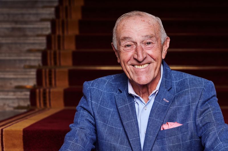 Len Goodman trägt einen blau karierten Blazer und ein rosa Einstecktuch und sitzt auf einer mit rotem Teppich ausgelegten Treppe